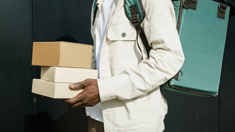 Close-up of a person holding three cardboard gift boxes, each wrapped in plain brown or white paper, with a black background. The individual's hand is visible, grasping the boxes, and they are dressed
