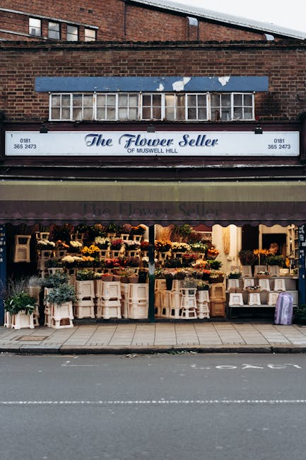 The exterior of The Flower Seller of Muswell Hill showcases a charming floral shop with large display windows filled with an array of vibrant fresh flowers arranged in bouquets and containers. The flo