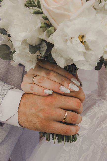 Close-up of a bride and groom's hands holding a bouquet of fresh flowers, featuring white lisianthus and green foliage, with the bride's manicured nails painted a pale silver and both wearing simple g
