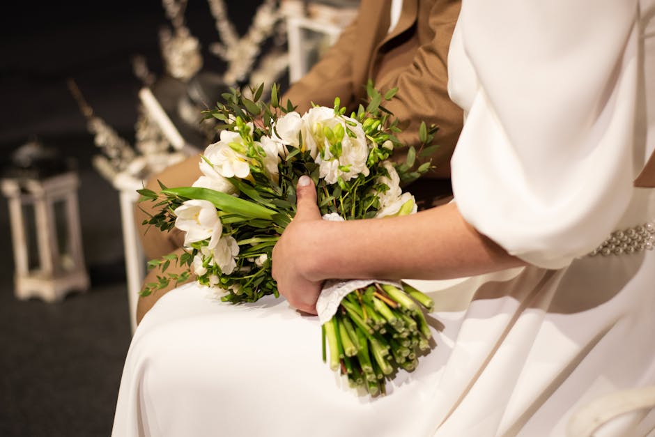 A bride holding a bouquet of fresh white flowers, including lisianthus and greenery, wrapped with a white lace ribbon, seated indoors with soft, warm lighting. Part of her white wedding dress with a p