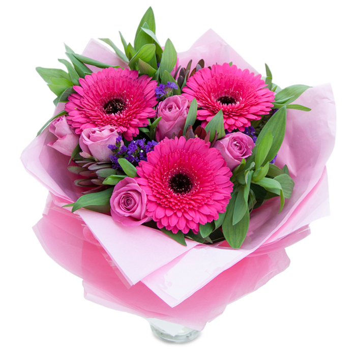 There is a playful elegance in this hand-tied bouquet: three vivid fuchsia Gerbera daisies claim the focal point with their fan-like petals and strong, dark centres that steady the composition. Paired alongside are several lavender-pink roses, their softly muted hue leaning toward dusty rose and their petals still retaining that new-bloom tightness that speaks of freshness. Between these main flowers, sprays of tiny purple statice add delicate punctuation marks of colour and a slightly papery texture that contrasts the velvet of the roses. Lush green foliage - likely salal or the rounded leaves of eucalyptus gathered from local suppliers around Gladstone Park and Dollis Hill - frames the arrangement and offers a natural, garden-like fullness.
The bouquet is wrapped as a gift: an inner sheet of matte pale pink tissue cushions and softens the visual base while an outer layer of slightly crinkled, translucent fuchsia-pink cellophane provides a subtle sheen and cohesive pink theme. At the lower edge the clear top rim of a glass vase is revealed, suggesting the flowers are ready to be displayed on a mantel or dining table. Imagine the tactile contrasts - the smoothness of rose petals, the crisp snap of foliage, the whisper of cellophane - and the scent, where roses give a gentle perfume and gerberas add a faint, green sweetness. This arrangement feels made for celebrations, quiet anniversaries, or as a thoughtful gesture that will brighten a home in Dollis Hill, Neasden, or nearby Willesden Green.